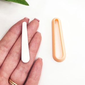 Könnte beinhalten: Eine weiße Plastikform zum Herstellen von Polymer Clay Ohrringen. Die Form ist lang, dünn und rechteckig mit abgerundeten Kanten. Die Form wird neben einem fertigen Polymer Clay Ohrring gezeigt.