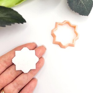 May include: A white, geometric tile held in a hand, next to a peach-coloured cookie cutter in the same shape. The tile has a starburst design. The cookie cutter is made of plastic. The background is white.