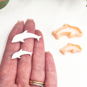 以下が含まれることがあります： 白いプラスチック製のイルカ型クッキーカッターが2つ、片方がもう片方より大きいものが、手で持たれています。オレンジ色のプラスチック製のイルカ型クッキーカッターが2つ、片方がもう片方より大きいものが、白い表面の上に置かれています。