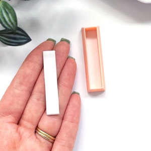 Könnte beinhalten: Ein weißer rechteckiger Aufkleber mit einer rosa rechteckigen Box. Der Aufkleber wird in einer Hand gehalten.