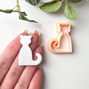 May include: White plastic cat cookie cutter with a detailed outline. The cutter is held in a hand, and a second, larger, peach-coloured cutter is visible in the background.