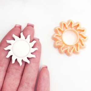 Op de afbeelding: Twee zonvormige koekjesvormen, een witte en een oranje. De witte vorm wordt in een hand gehouden, terwijl de oranje vorm op een wit oppervlak ligt.
