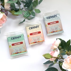 May include: Three packets of Cernit polymer clay in green, beige, and blue. Each packet is labelled "NATURE" with text in multiple languages. The clay is packaged in clear plastic, with a white background and floral accents.