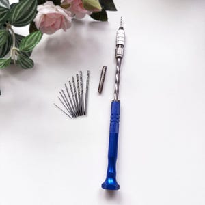 May include: A manual pin vice hand drill featuring a bright blue knurled handle and a silver twisted metal shaft. The tool includes a silver collet and a set of ten graduated metal drill bits arranged in a fan shape on a plain white surface.