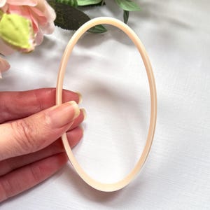 Op de afbeelding: Een licht perzikvormig, ovaalvormig object. Het object wordt door een hand vastgehouden, met een zachte achtergrond van roze en groene bloemelementen. Het object lijkt gemaakt te zijn van een glad, mogelijk plastic materiaal.