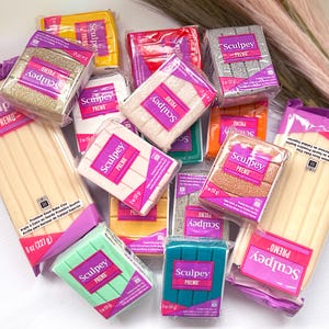 May include: An assortment of Sculpey Premo brand oven-bake clay in various colours, including gold, pink, green, blue, grey, and orange. Each block of clay is labelled with the brand name and the words "Premo" and "Oven-Bake Clay".