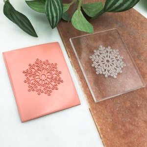 Puede incluir: Un sello de acrílico transparente con un diseño de copo de nieve. El sello está sobre una lámina de goma rosa.