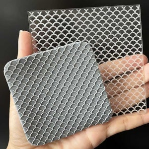 Op de afbeelding: Een grijze, vierkante siliconen mal en een heldere acrylplaat, beide met een herhalend visschubbenpatroon. De mal wordt in een hand gehouden, wat de gedetailleerde textuur laat zien. De acrylplaat is transparant.