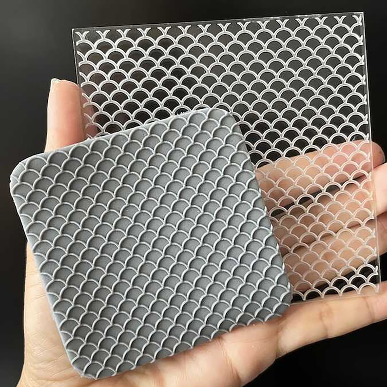Op de afbeelding: Een grijze, vierkante siliconen mal en een heldere acrylplaat, beide met een herhalend visschubbenpatroon. De mal wordt in een hand gehouden, wat de gedetailleerde textuur laat zien. De acrylplaat is transparant.