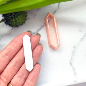 May include: A white, rectangular, flat, geometric shape with rounded edges. The shape is being held in a hand. A pink, rectangular, geometric shape with rounded edges is also shown. The shape is similar to the white shape but is slightly larger and has a hollow center.