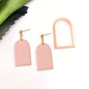 May include: A pair of blush pink dangle earrings with gold-colored hardware. The earrings have a rounded top and a rectangular shape. A matching arch-shaped cutter is also present.
