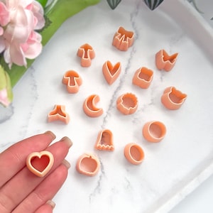 May include: A set of 14 pink plastic cookie cutters in various shapes, including a heart, a mushroom, a crescent moon, a pumpkin, a ghost, a bow, a butterfly, a cat, a flower, a circle, and a square.