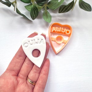 May include: White plastic Ouija board cookie cutter with a heart shape and the word "OUIJA" embossed on the front. A matching pink plastic cookie cutter is shown in the background.