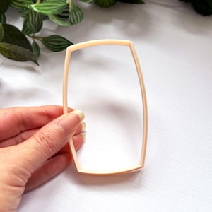 May include: A light peach-colored cookie cutter with a rectangular shape featuring slightly curved sides. The frame is constructed with a thin, raised edge, creating a hollow center for cutting dough into a specific geometric form.