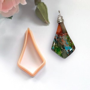 May include: A pendant featuring a green, blue, and orange gemstone with copper flecks. The gemstone is set in a silver wire wrap and hangs from a silver bail. The pendant is displayed next to a peach-coloured plastic mould.