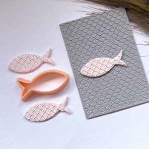 May include: A set of three pink fish-shaped cookie cutters with a scale pattern. The cutters are on a white surface next to a gray silicone mat with a scale pattern.