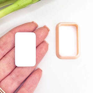Può includere: Un pezzo rettangolare bianco solido con angoli arrotondati e una cornice rettangolare separata color pesca con bordi arrotondati. Il pezzo bianco viene tenuto in mano, mentre la cornice color pesca è posizionata accanto su una superficie bianca.