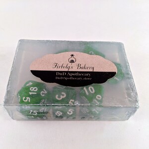 May include: A set of green dice with white numbers inside a clear, rectangular container. The dice are for tabletop games. The container has a label that reads "Firbolg's Bakery" and "DnD Apothecary".