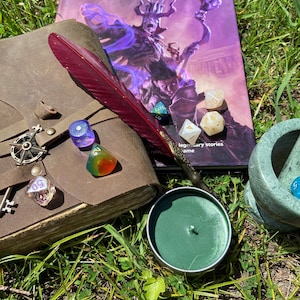 May include: A collection of tabletop gaming accessories on grass. Includes a leather-bound journal, a red feather quill, dice, a green candle, and a Dungeon Master's Guide book. The book cover features a fantasy illustration with a purple and blue color scheme.