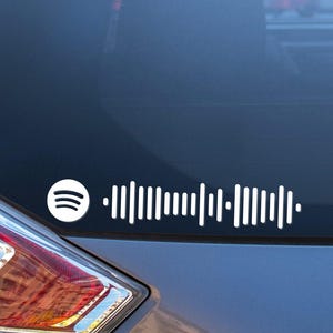 May include: White Spotify music code decal on a car's rear window. The decal features the Spotify logo and a series of vertical lines representing a music track's waveform. The car is a dark gray color.