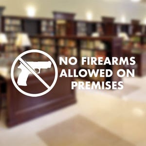 May include: A white sign with a crossed-out handgun symbol and the text "NO FIREARMS ALLOWED ON PREMISES." The sign is in front of a blurred background of a library or office setting.