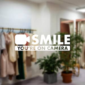 May include: White text on a blurred background of a clothing store. The text reads "SMILE YOU'RE ON CAMERA" with a camera icon. Clothing racks with various garments and potted plants are visible.