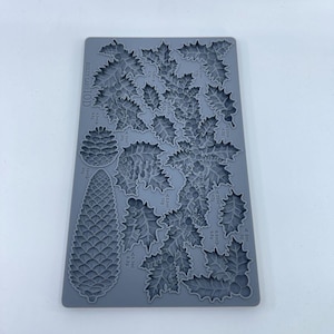 Op de afbeelding: Een grijze siliconen mal met hulstbladeren en dennenappels. De mal heeft de tekst "HOLLY LANE LTD" linksboven. Het is ontworpen voor het maken van decoratieve elementen voor knutsel- en bakprojecten.