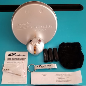 May include: A silver and chrome car gear shift knob with a gold accent ring. The knob is on a white background with a silver round container with the text "Ichibahn" in black and Japanese characters. The image also includes a black velvet bag, a silver bottle opener with a keychain, and a small instruction sheet.