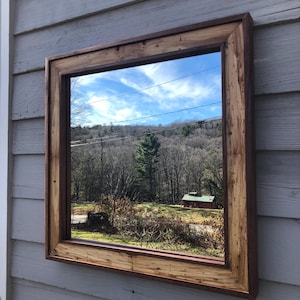 May include: A large square mirror with a rustic wooden frame. The frame is made of reclaimed wood with a natural finish. The mirror reflects a view of a wooded area with a small house in the distance.