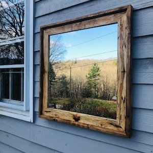 May include: A rustic wooden framed mirror with a natural wood finish. The mirror reflects a view of a forest and a distant hillside.