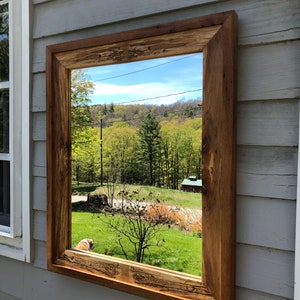 May include: A rustic wooden mirror with a thick frame, reflecting a view of a green forest and a blue sky. The mirror is mounted on a gray wooden wall.