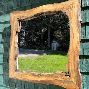 May include: A square mirror featuring a natural live edge wooden frame with visible bark, dark grain patterns, and light brown wood tones. The frame has an irregular, organic shape with rough, textured edges surrounding the reflective glass surface.