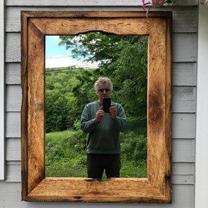May include: A rustic wooden mirror with a thick, natural wood frame. The mirror reflects a man holding a smartphone in front of a green, leafy background.