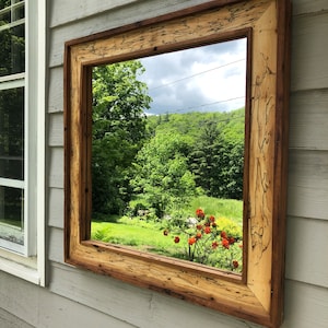May include: A rectangular mirror with a rustic wooden frame. The frame is made of reclaimed wood with a natural wood finish. The mirror reflects a view of a green forest and a blue sky with white clouds.