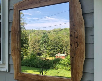 May include: A rectangular mirror with a natural wood frame. The frame has a live edge design, showcasing the wood's organic shape and texture. The mirror reflects a scenic outdoor view of trees, a red building, and a blue sky.