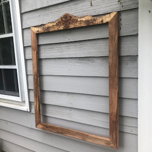 May include: A rustic wooden frame with a unique, irregular top edge. The frame is made of natural wood with a warm brown color and is mounted on a gray wooden wall.