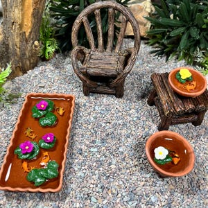 May include: A miniature fairy garden scene with a wooden chair, a small table, and three ponds with pink and white flowers and goldfish. The ponds are made of terracotta and are surrounded by small pebbles.