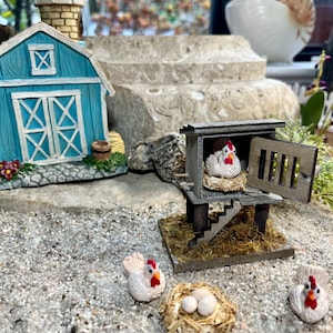 Puede incluir: Un gallinero en miniatura de madera con una gallina blanca en el interior, junto con tres gallinas blancas y un nido de huevos en el exterior. El gallinero est&aacute; hecho de madera y tiene una peque&ntilde;a puerta. Las gallinas son todas blancas con crestas rojas. El nido est&aacute; hecho de paja y tiene tres huevos blancos en su interior.