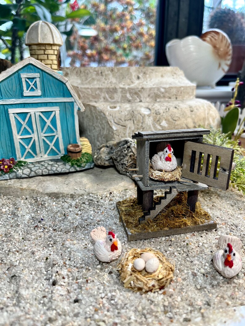 Puede incluir: Un gallinero en miniatura de madera con una gallina blanca en el interior, junto con tres gallinas blancas y un nido de huevos en el exterior. El gallinero est&aacute; hecho de madera y tiene una peque&ntilde;a puerta. Las gallinas son todas blancas con crestas rojas. El nido est&aacute; hecho de paja y tiene tres huevos blancos en su interior.