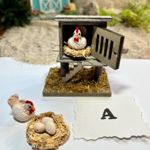 Puede incluir: Un gallinero de madera en miniatura con una gallina blanca dentro y una gallina blanca fuera con un nido de tres huevos blancos. El gallinero tiene una peque&ntilde;a escalera de madera y una puerta abierta. El gallinero est&aacute; sentado sobre una cama de paja. Hay una tarjeta blanca con la letra A en ella.