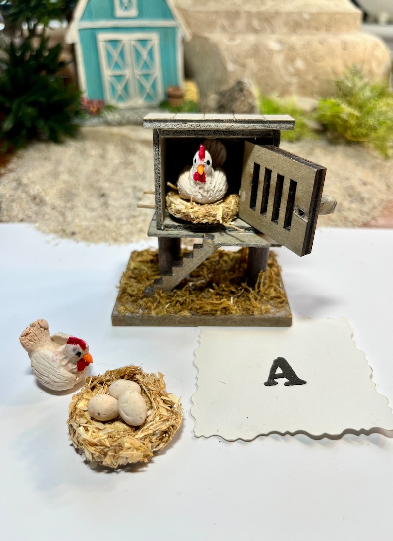 Puede incluir: Un gallinero de madera en miniatura con una gallina blanca dentro y una gallina blanca fuera con un nido de tres huevos blancos. El gallinero tiene una peque&ntilde;a escalera de madera y una puerta abierta. El gallinero est&aacute; sentado sobre una cama de paja. Hay una tarjeta blanca con la letra A en ella.