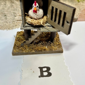 Puede incluir: Un gallinero de madera en miniatura con una gallina blanca en su interior. El gallinero tiene una peque&ntilde;a puerta que est&aacute; abierta y una escalera que lleva hasta el gallinero. El gallinero est&aacute; sentado sobre una cama de paja marr&oacute;n. La imagen tiene la letra "B" en una tarjeta blanca debajo del gallinero.