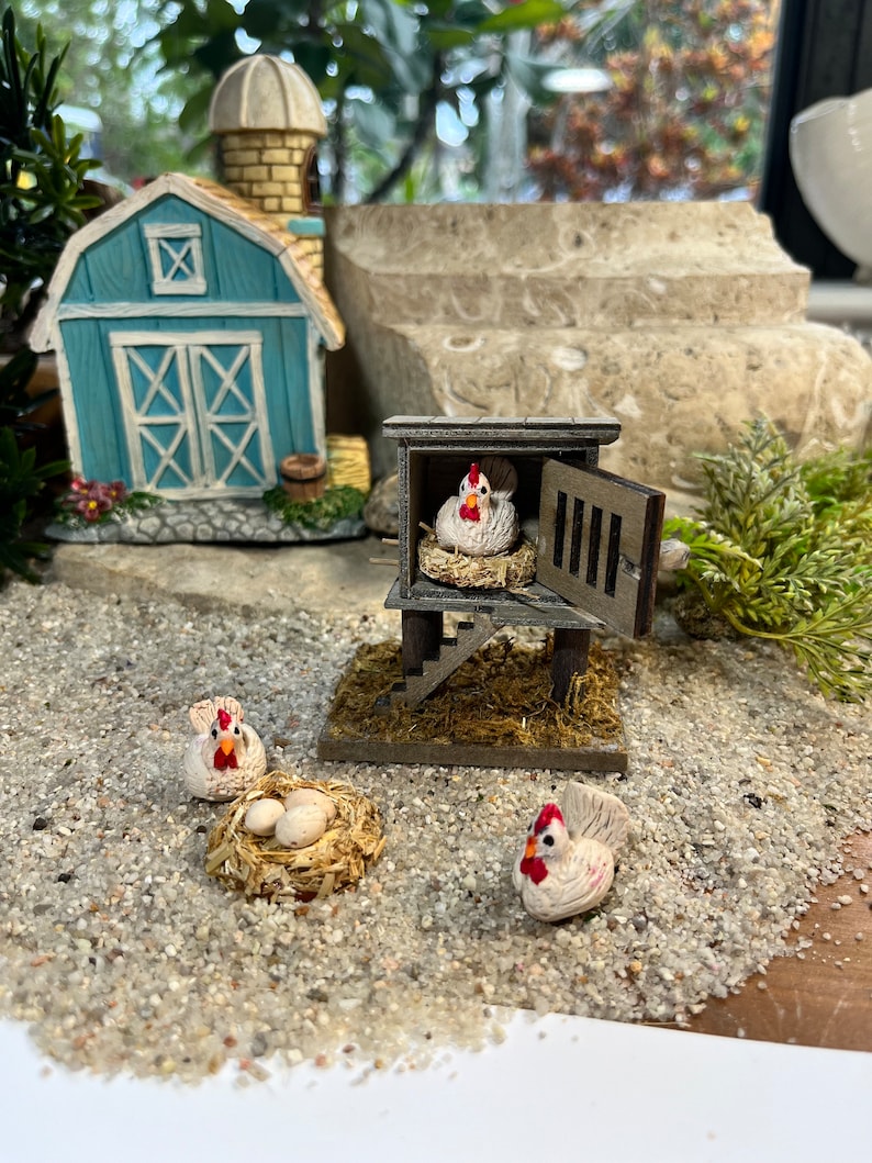 Puede incluir: Un gallinero de madera en miniatura con una gallina blanca dentro y dos gallinas blancas afuera. El gallinero tiene una peque&ntilde;a puerta de madera y una escalera. Hay tres huevos blancos en un nido fuera del gallinero.