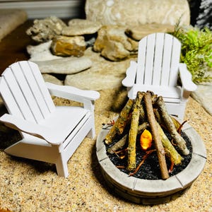May include: Miniature outdoor scene with two white Adirondack chairs and a stone fire pit. The fire pit contains logs and a glowing orange light. The scene is set on a sandy surface with rocks in the background.