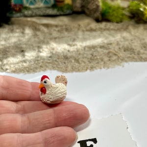 Puede incluir: Una peque&ntilde;a figurita de pollo de cer&aacute;mica blanca con una cresta roja y un pico naranja. El pollo est&aacute; sentado en una mano, y hay una tarjeta blanca con la letra 'E' impresa en tinta negra.