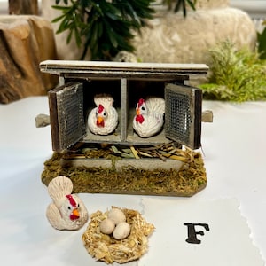 Puede incluir: Un gallinero de madera en miniatura con dos gallinas blancas dentro y una gallina blanca y tres huevos blancos en un nido afuera. El gallinero tiene una puerta de madera marr&oacute;n y una ventana de malla met&aacute;lica. La letra F est&aacute; escrita en un trozo de papel blanco.