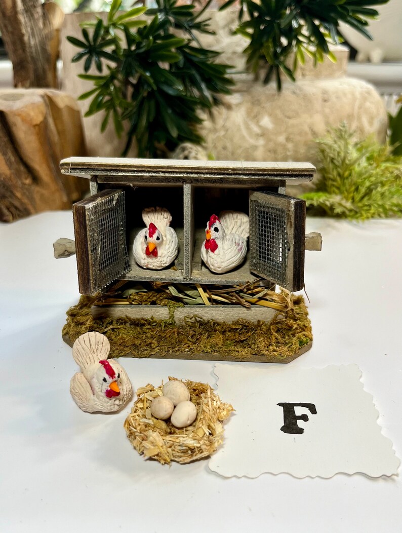 Puede incluir: Un gallinero de madera en miniatura con dos gallinas blancas dentro y una gallina blanca y tres huevos blancos en un nido afuera. El gallinero tiene una puerta de madera marr&oacute;n y una ventana de malla met&aacute;lica. La letra F est&aacute; escrita en un trozo de papel blanco.