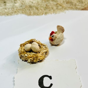 Puede incluir: Una figurita de pollo de cer&aacute;mica blanca con una cresta roja y un nido con dos huevos blancos. El nido est&aacute; hecho de paja y est&aacute; sobre una superficie blanca. La letra "C" est&aacute; escrita en una tarjeta blanca con un borde festoneado.