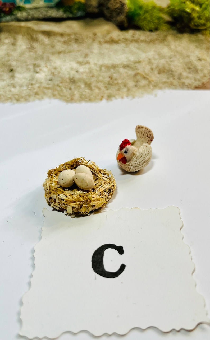 Puede incluir: Una figurita de pollo de cer&aacute;mica blanca con una cresta roja y un nido con dos huevos blancos. El nido est&aacute; hecho de paja y est&aacute; sobre una superficie blanca. La letra "C" est&aacute; escrita en una tarjeta blanca con un borde festoneado.