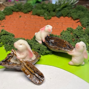 May include: Three white ceramic bunnies with pink ears, two are in brown boats with oars, one is pushing a brown boat.
